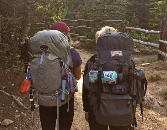 2 female backpackers headed up a wooded trail, both carrying large packs with camping items hanging from straps