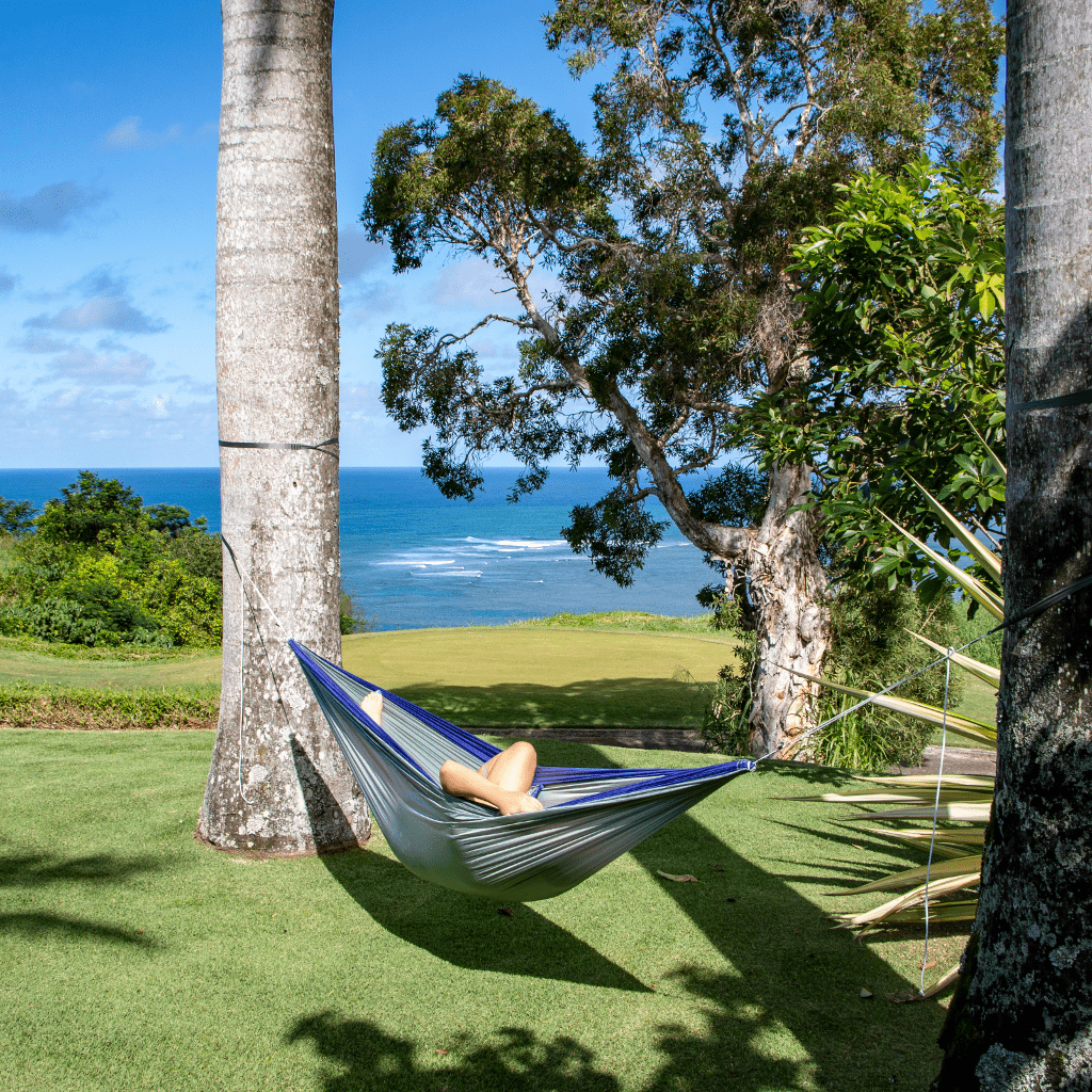 Person relaxing in a Hummingbird Hammocks Ultralight Double Hammock between palm trees, overlooking a scenic ocean view with lush greenery around.