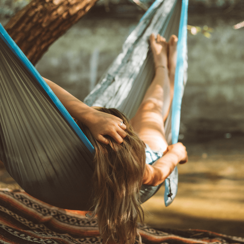 A person relaxing in a Hummingbird Hammocks ultralight double hammock, their arm stretching out and head resting back, surrounded by a sunlit natural setting.