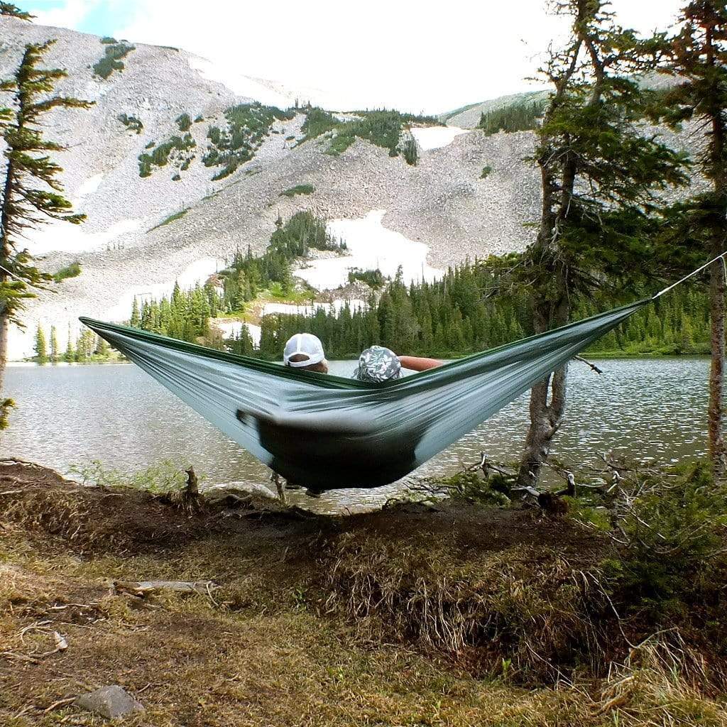 Person relaxing in a Hummingbird Hammocks Ultralight Double Hammock between two trees with a scenic view of a mountain lake and forest in the background.
