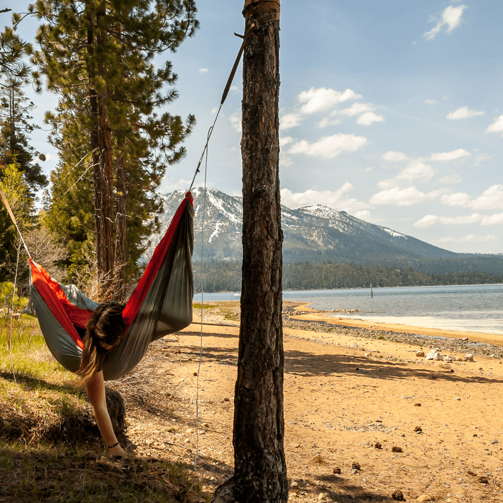 Person relaxing in a Hummingbird Hammocks Ultralight Double Hammock strung between two trees by a lake with a mountainous backdrop.