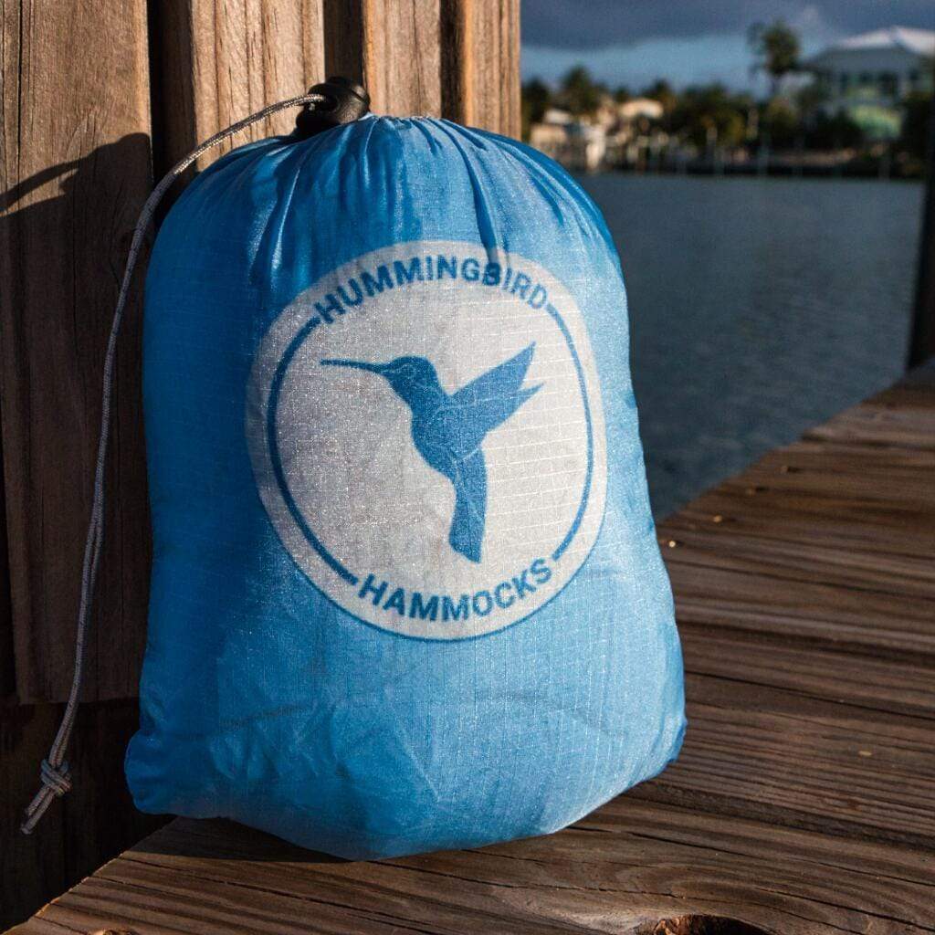A blue drawstring bag labeled "Hummingbird Hammocks" with a logo of a hummingbird, resting on a wooden dock beside water, featuring an Ultralight Long Hammock design.
