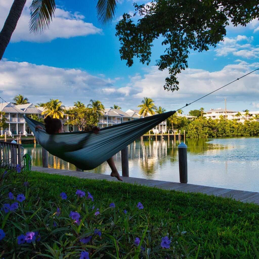 A person relaxing in a Hummingbird Hammocks ultralight long hammock by a calm waterfront, surrounded by lush greenery and blue skies.