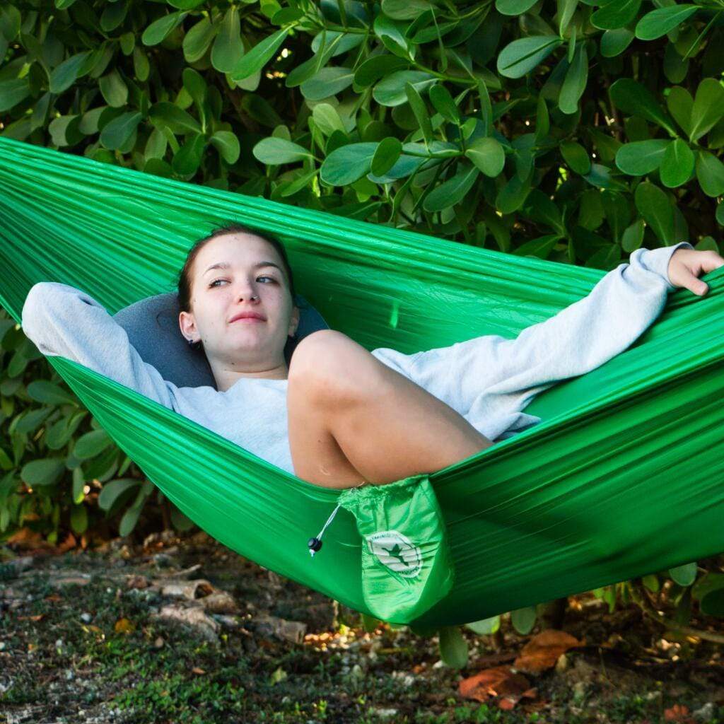 A person lounging in an ultralight Hummingbird Hammocks long hammock among lush green foliage, looking relaxed and staring directly at the camera.