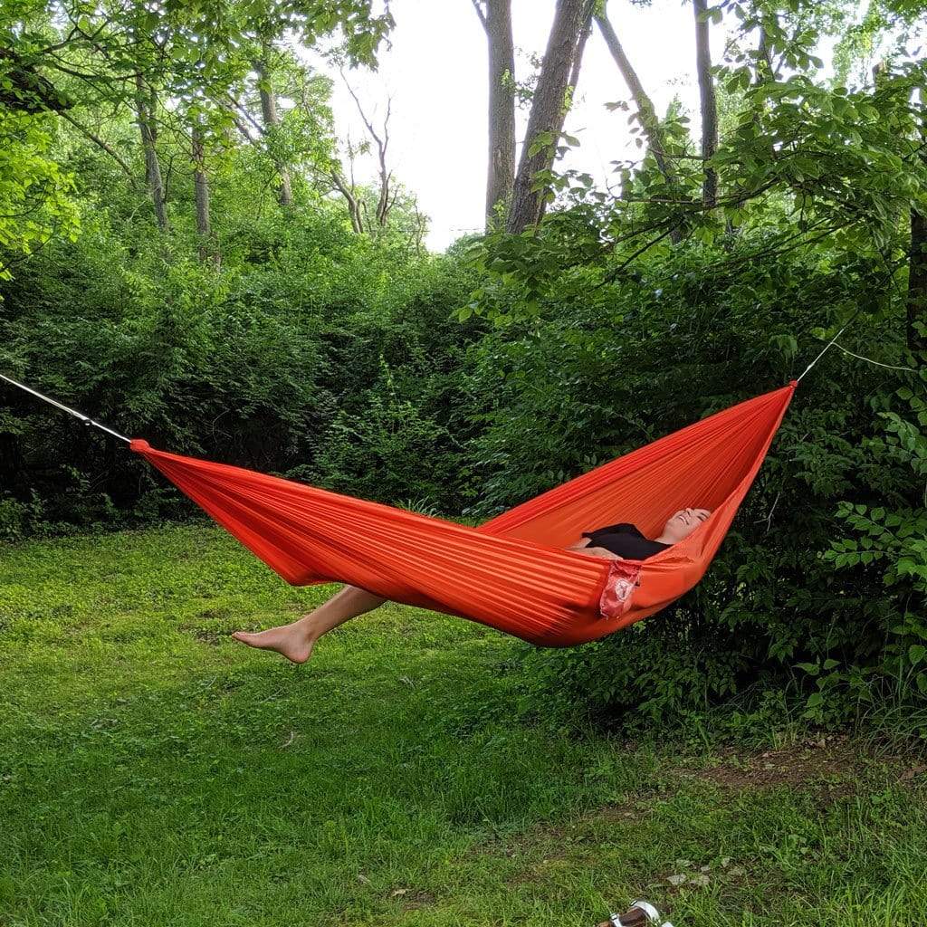A person lounging in a Hummingbird Hammocks ultralight long hammock strung between trees in a lush green backyard.