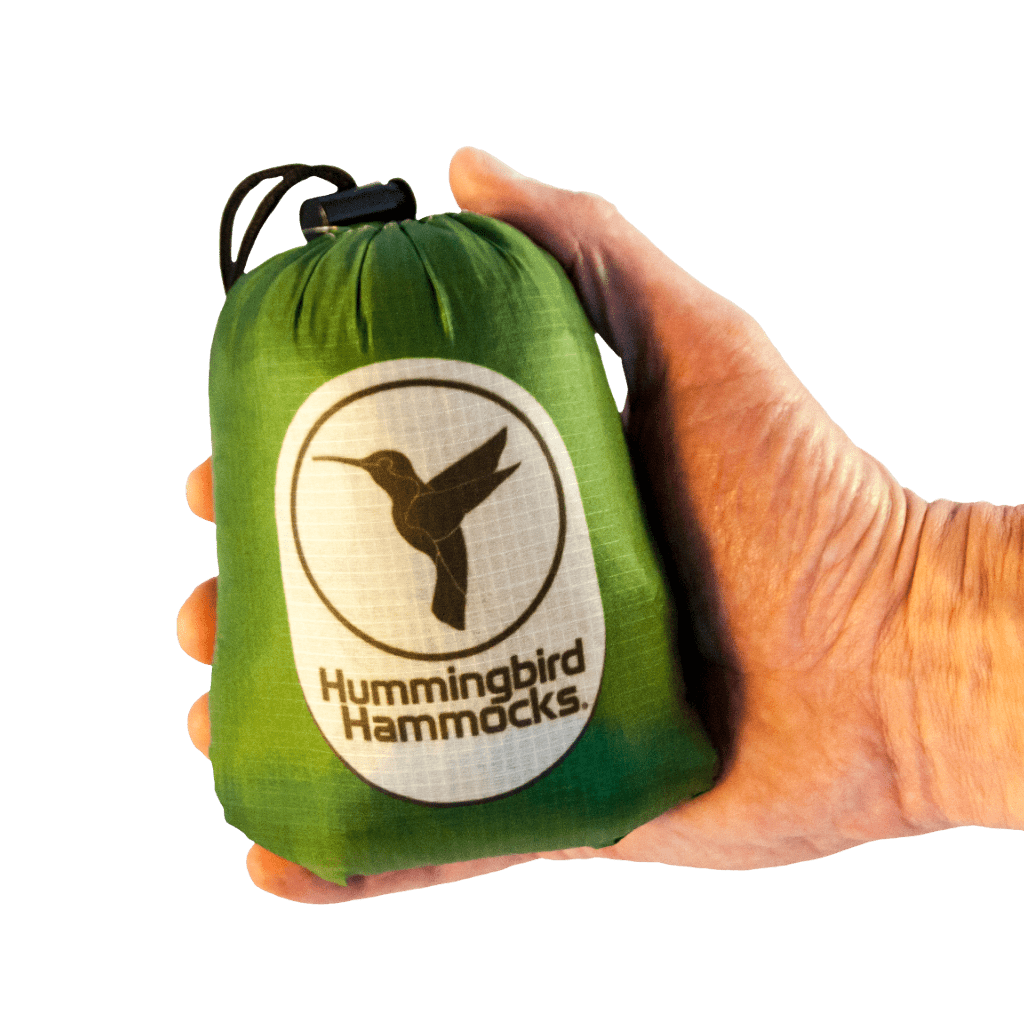 A hand holding a small green stuff sack with the Button Link attachment system, featuring the logo "Hummingbird Hammocks" with a silhouette of a hummingbird containing the Single+ Hammock.