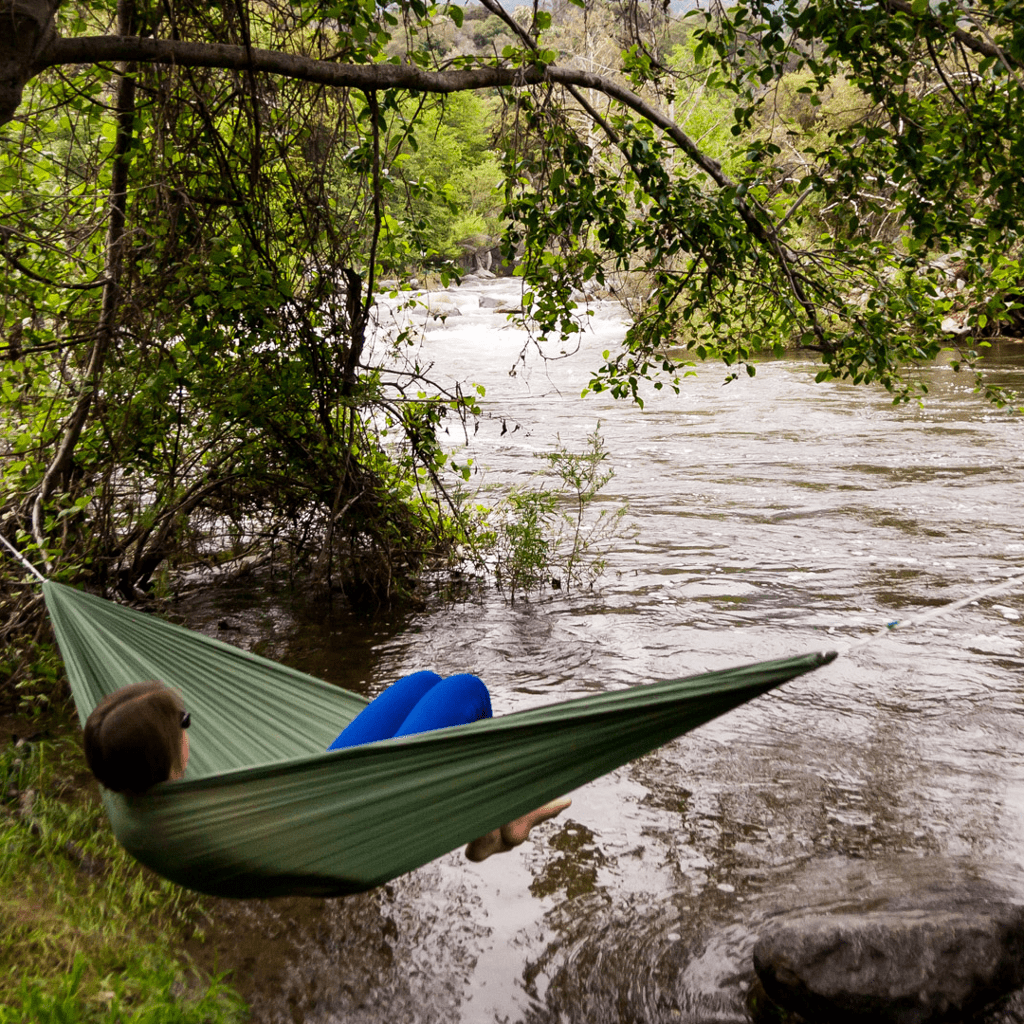 A person relaxes in a green Hummingbird Hammocks Ultralight Single+ Hammock over a river, surrounded by lush greenery and rocks, with rapids in the background.