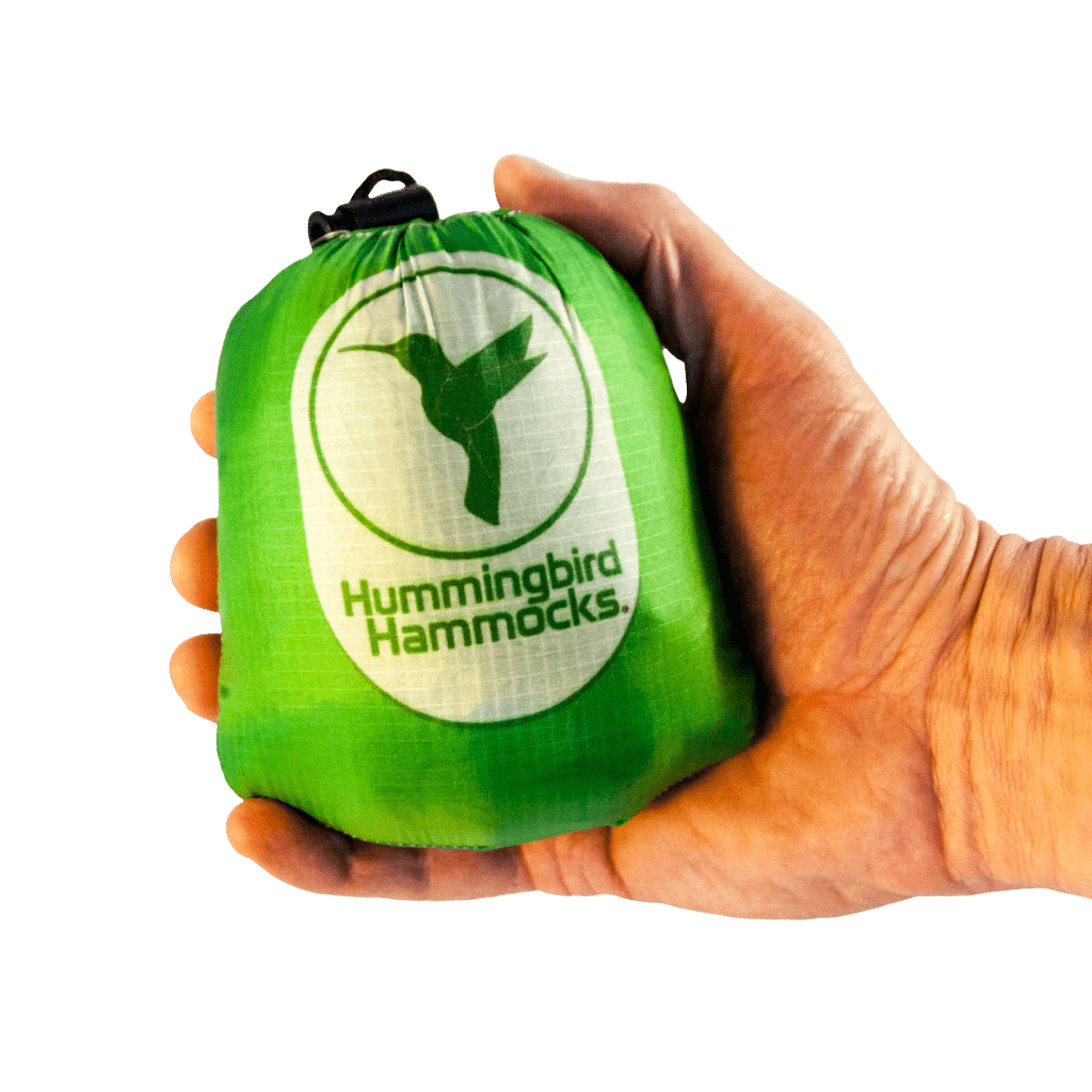 A hand holding a small, green drawstring bag with the "Hummingbird Hammocks" logo featuring a white hummingbird silhouette and utilizing advanced parachute technology.