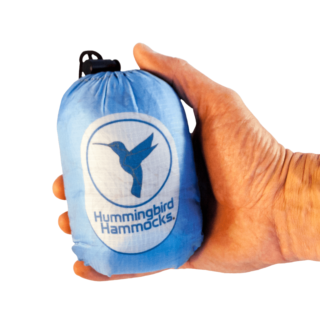 A hand holding a compact, blue Hummingbird Hammocks Single+ Hammock stuff sack with Button Link attachment system against a green background.