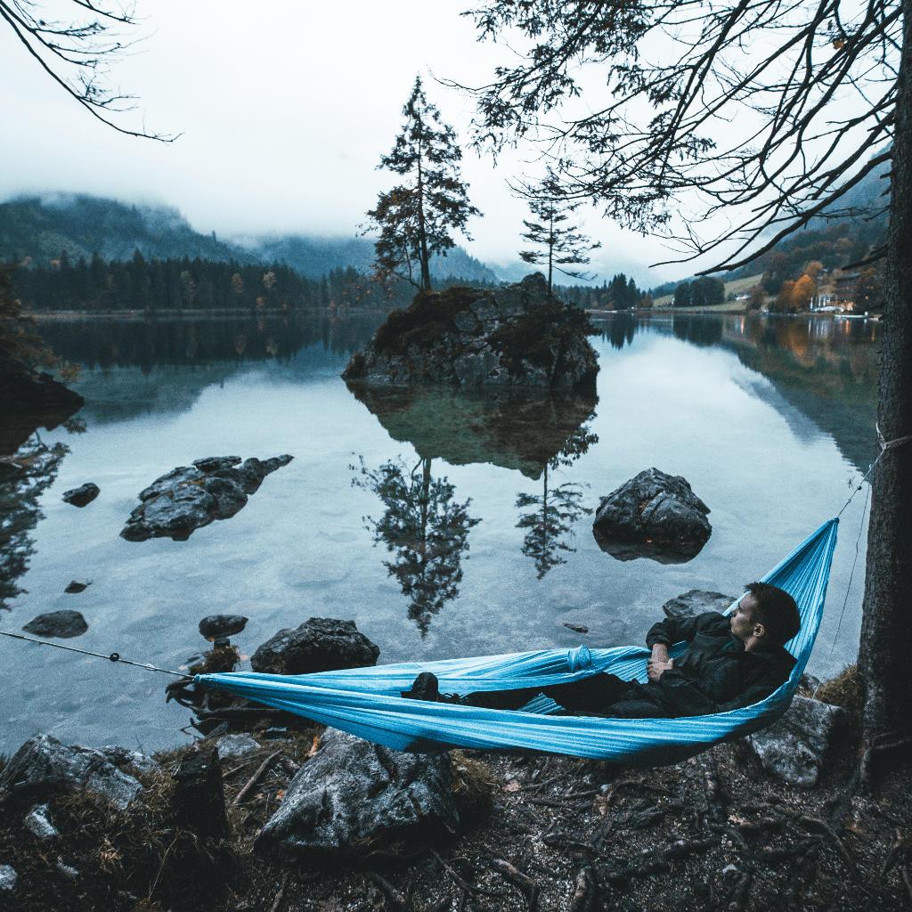 A person relaxing in a blue Ultralight Single+ Hammock by Hummingbird Hammocks suspended between trees by a tranquil, misty lake with scattered boulders and a forested background.