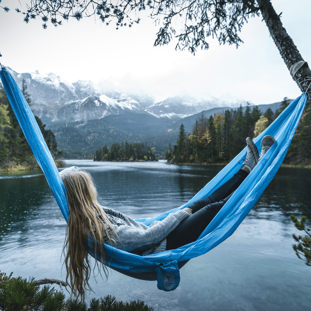 A person relaxing in a Hummingbird Hammocks Ultralight Single+ Hammock by a lake, gazing at snowy mountains in the background.