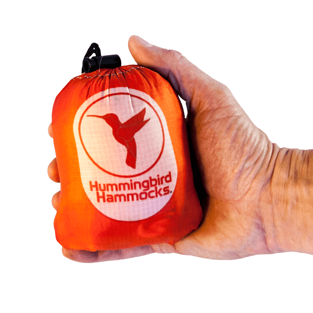 A hand holding a compact, orange stuff sack labeled "Hummingbird Hammocks Single+ Hammock," featuring parachute technology. The sack has a white logo of a hummingbird and a drawstring closure.