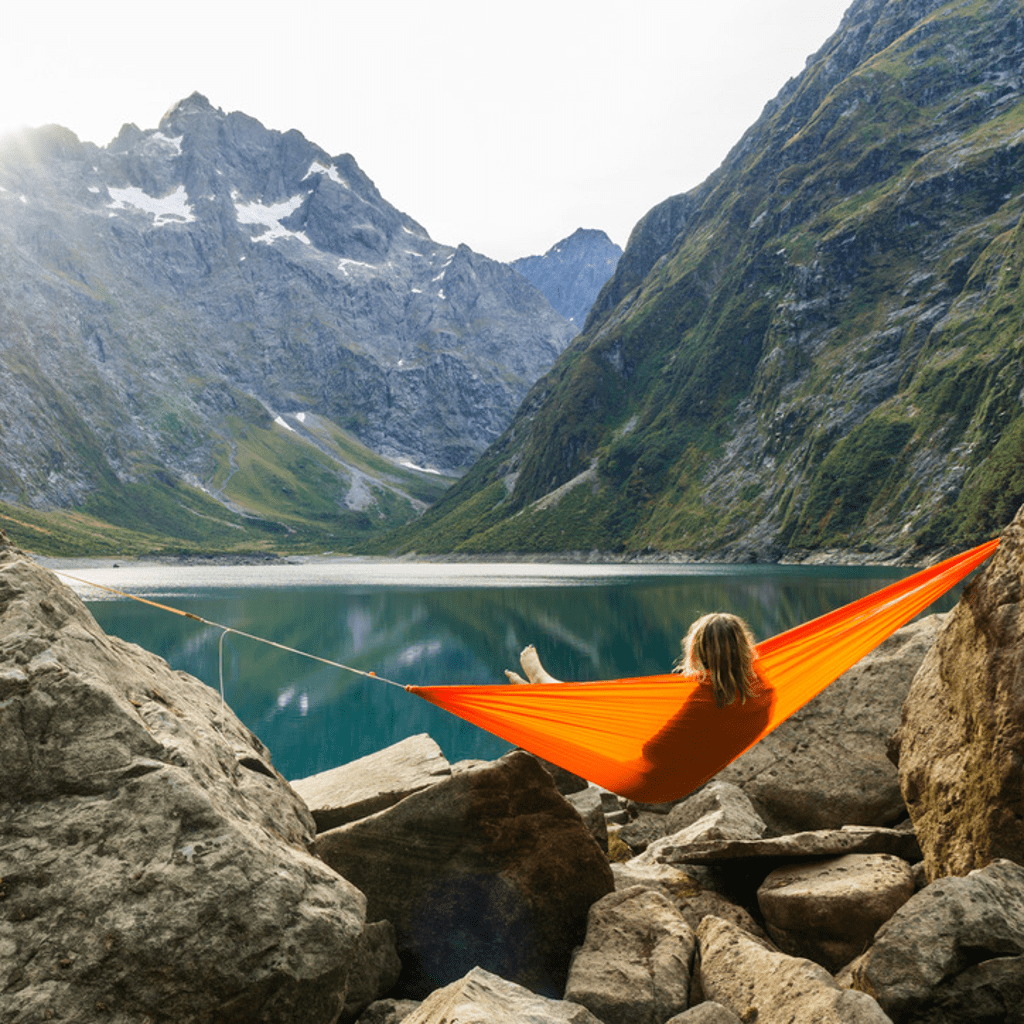 A person relaxes in a bright orange Hummingbird Hammocks Ultralight Single+ Hammock suspended between rocks, overlooking a serene mountain lake.