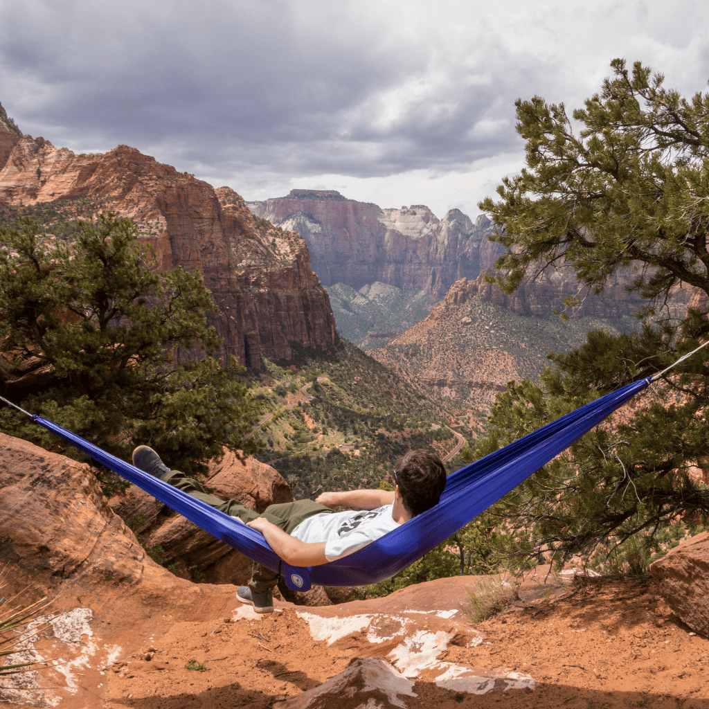 Person relaxing in a Hummingbird Hammocks Single+ hammock, made with parachute technology, overlooking a scenic canyon with lush greenery and cloudy skies.