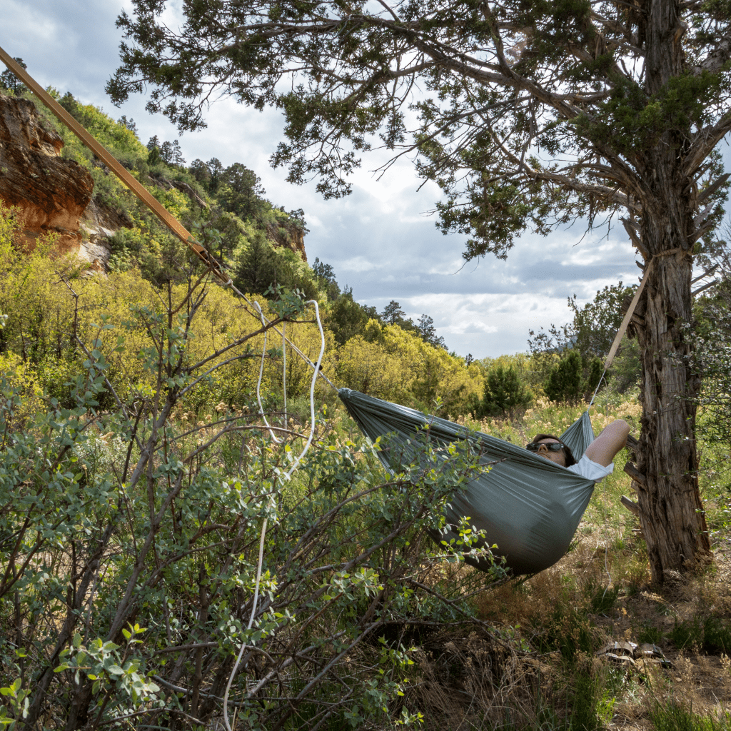 Person relaxing in a Hummingbird Hammocks Ultralight Single+ Hammock strung between trees in a lush, hilly area, with greenery and partly cloudy skies.