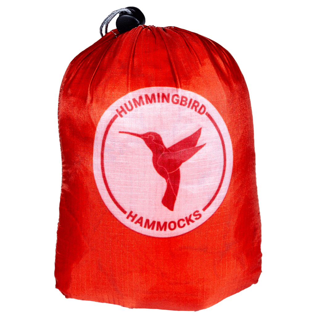 Red storage bag with "Hummingbird Hammocks" logo featuring a hummingbird graphic, isolated on a green background, includes an Ultralight Long Hammock from Hummingbird Hammocks.