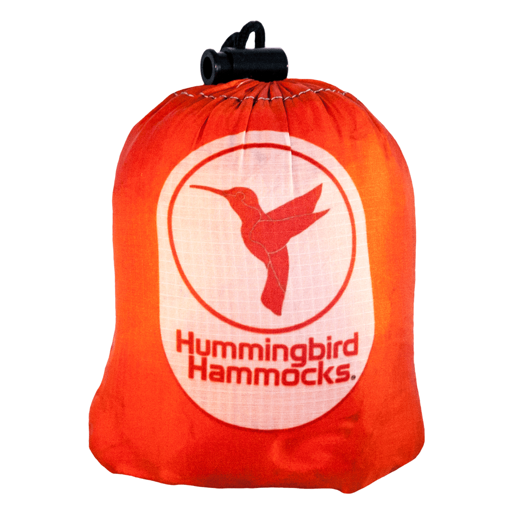 A bright orange stuff sack featuring the logo of Hummingbird Hammocks printed in white, designed with parachute technology, and displaying a hummingbird silhouette for the Single+ Hammock.