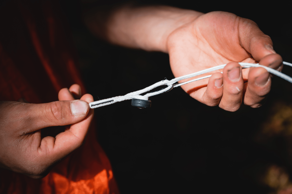 Two hands tying a knot in a white Spectra cord with a black toggle, set against a dimly lit background using the Hummingbird Hammocks Replacement Button Link Set.