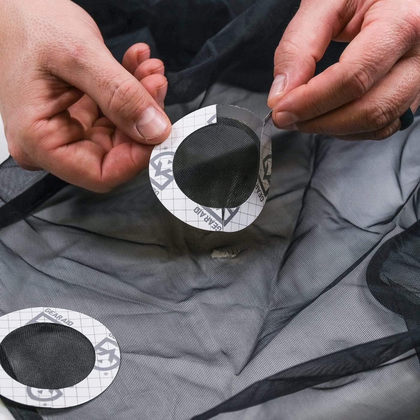 Hands applying a circular Tenacious Tape Mesh Patch from Hummingbird Hammocks to mend a tear in black mesh fabric.