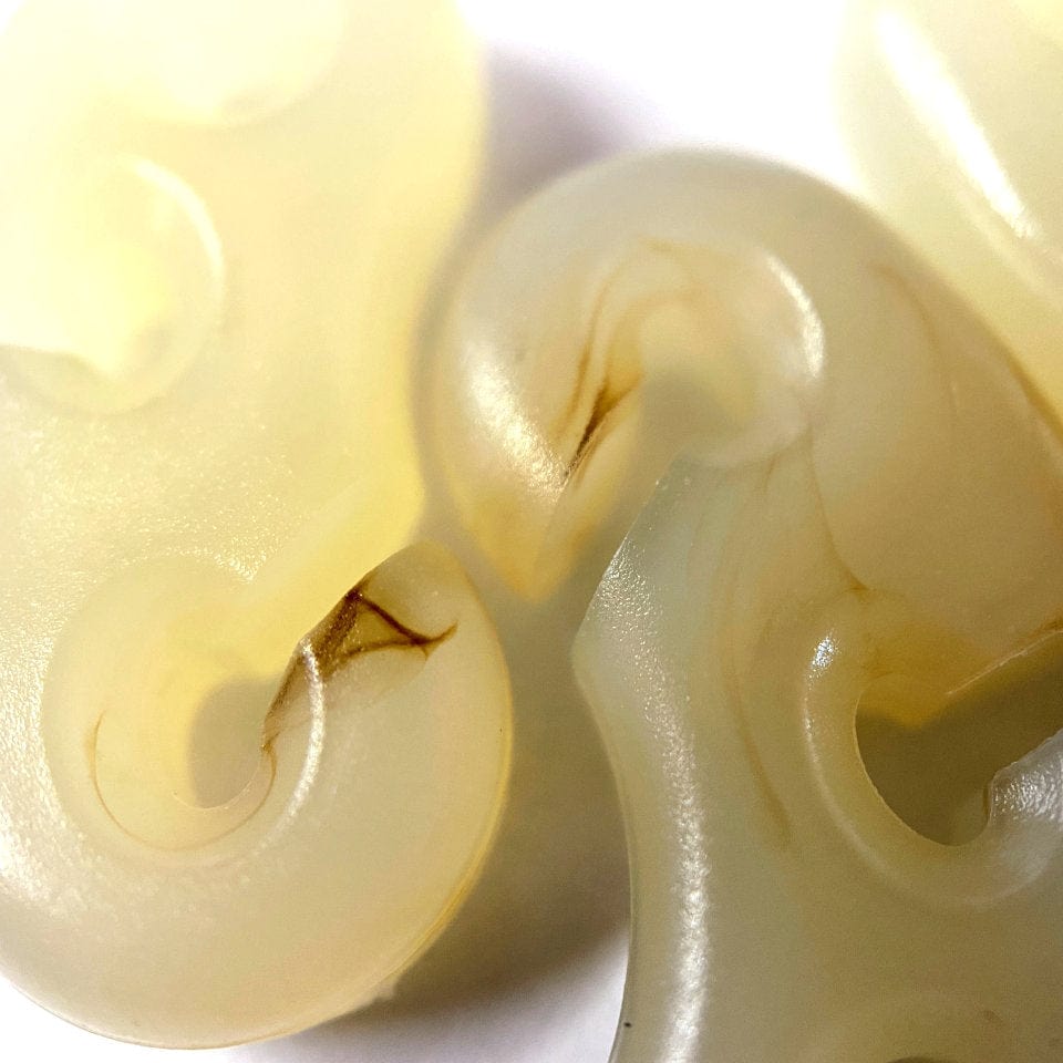 Close-up of translucent, amber inclusions inside off-white, curvy resin pieces displaying organic swirls and textures from injection molding - Glow Friction Adjusters from Hummingbird Hammocks.
