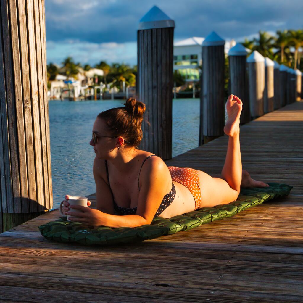 A woman in a bikini lies on a Junco Sleeping Pad by Hummingbird Hammocks on a wooden dock, holding a cup, with waterfront homes in the background during sunset.