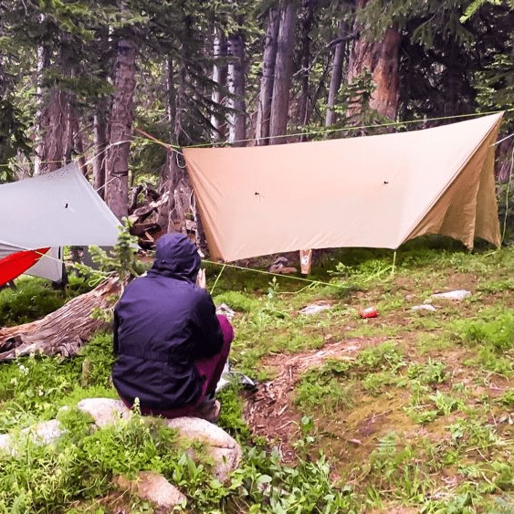 A person in a dark jacket sitting on a rock near a makeshift camping setup with a Pelican Rain Tarp by Hummingbird Hammocks in a dense forest.