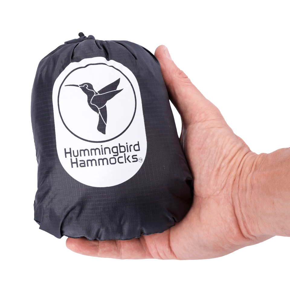 A hand holding a compact, gray Hummingbird Hammocks bag with a logo featuring a white hummingbird on a green background, ideal for carrying the Warbler Bug Net.