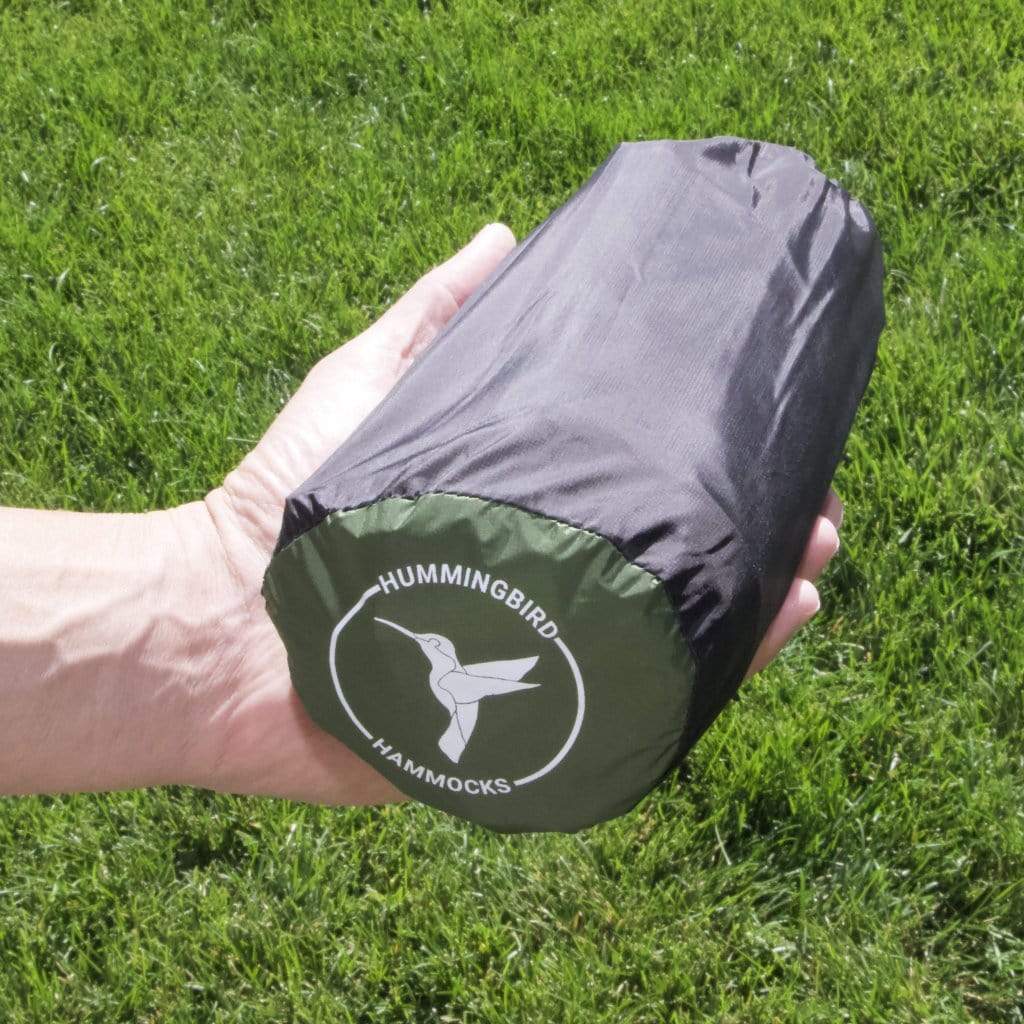 A hand holding a compact, rolled-up Junco Sleeping Pad bag on a green grass background from Hummingbird Hammocks.
