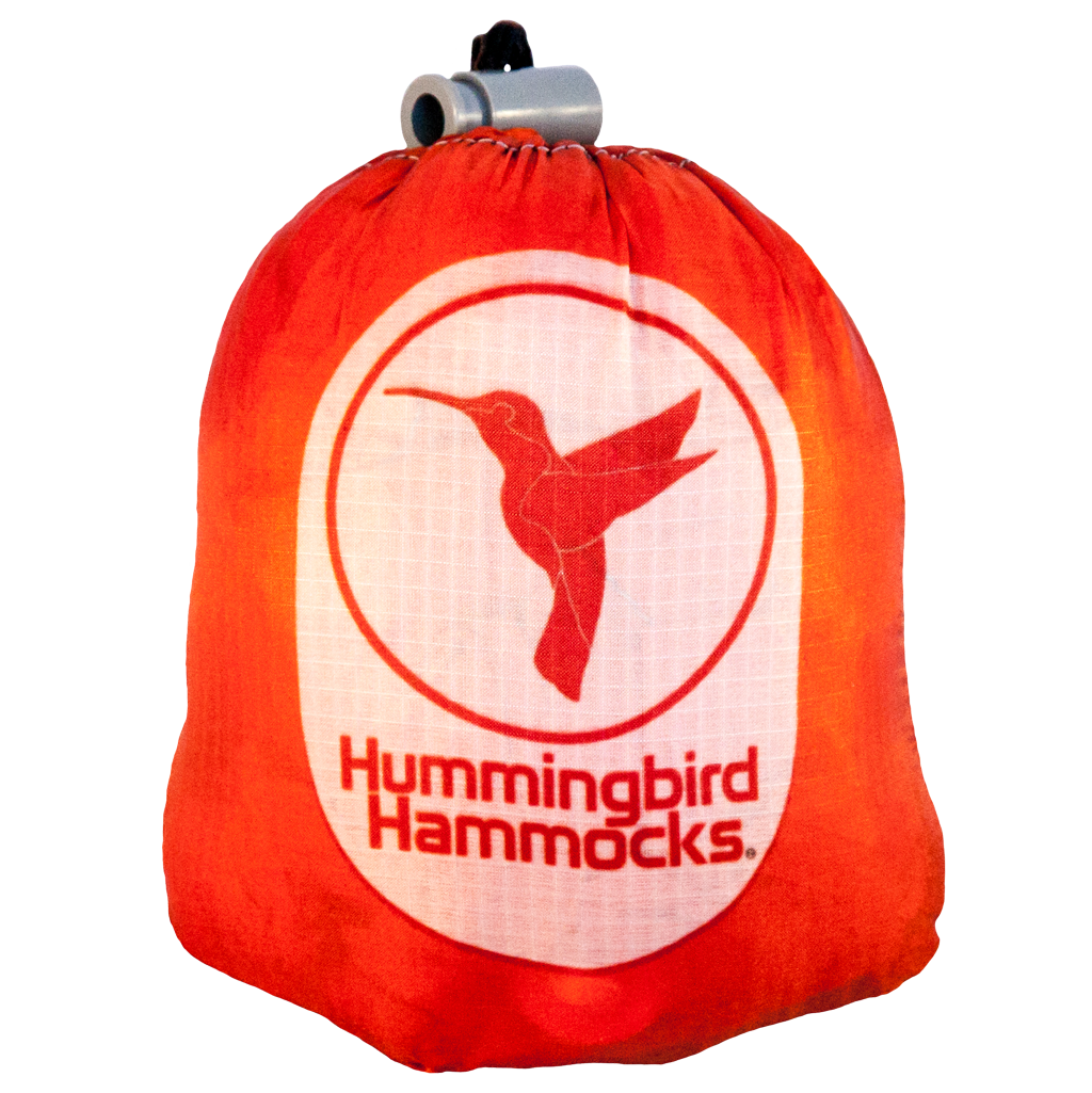 The Single Hammock by Hummingbird Hammocks comes in a red bag adorned with a white circle featuring a hummingbird logo and the text "Hummingbird Hammocks." It includes a gray closure mechanism at the top and highlights an innovative Button Link attachment system, ensuring effortless setup and convenience.