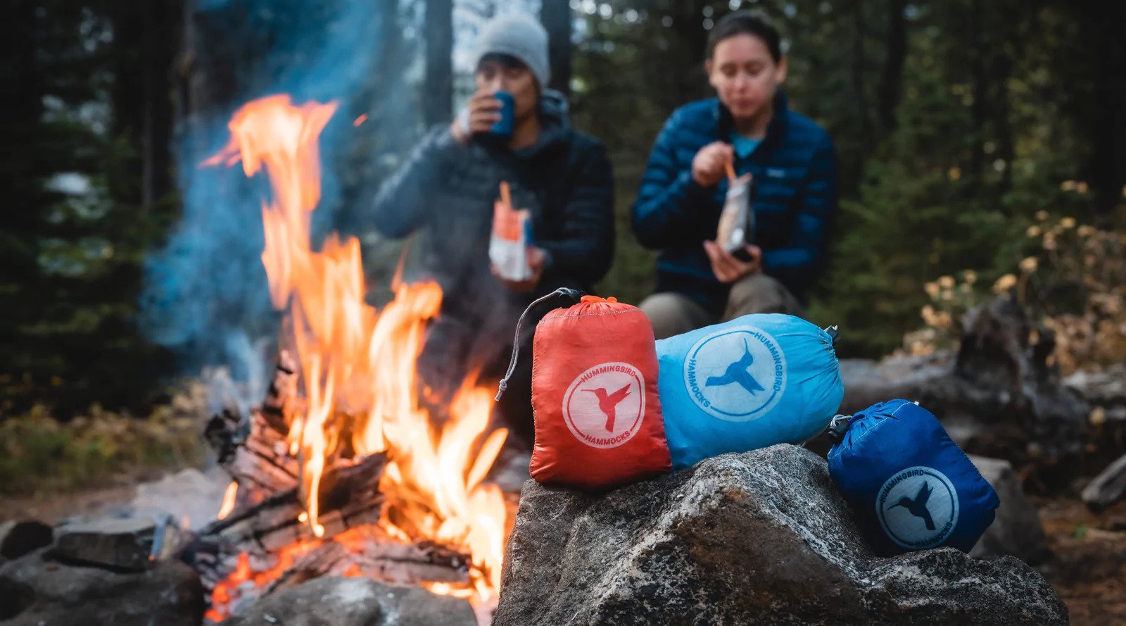 Three ultralight Single Plus Hammocks sitting nearby a campfire in the forest with people in the background eating a meal