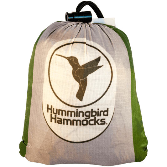 A drawstring backpack with the "Hummingbird Hammocks" logo featuring a silhouette of a hummingbird in flight, set against a white and green background, designed by an FAA Certified Parachute Rigger.