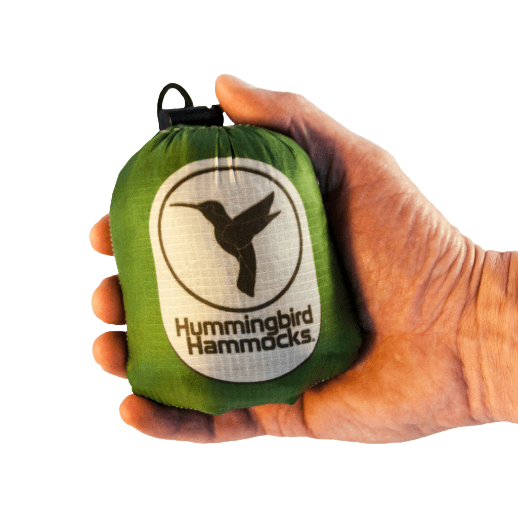 A hand holds a compact green Ultralight Hummingbird Hammocks Single Hammock bag with a white logo featuring a hummingbird.