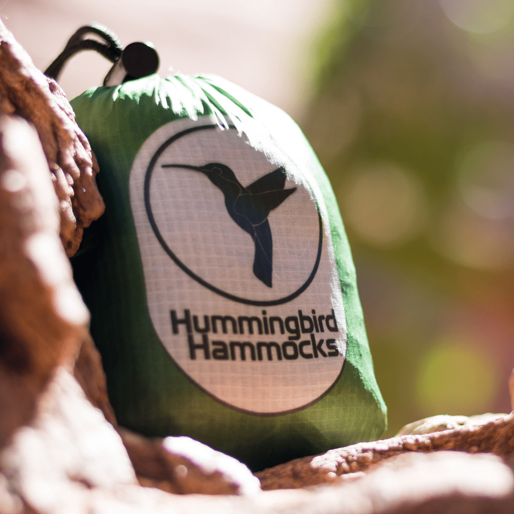 A close-up of a green Hummingbird Hammocks Ultralight Single Hammock bag with the company's logo, a hummingbird, poised on a rock in natural light.