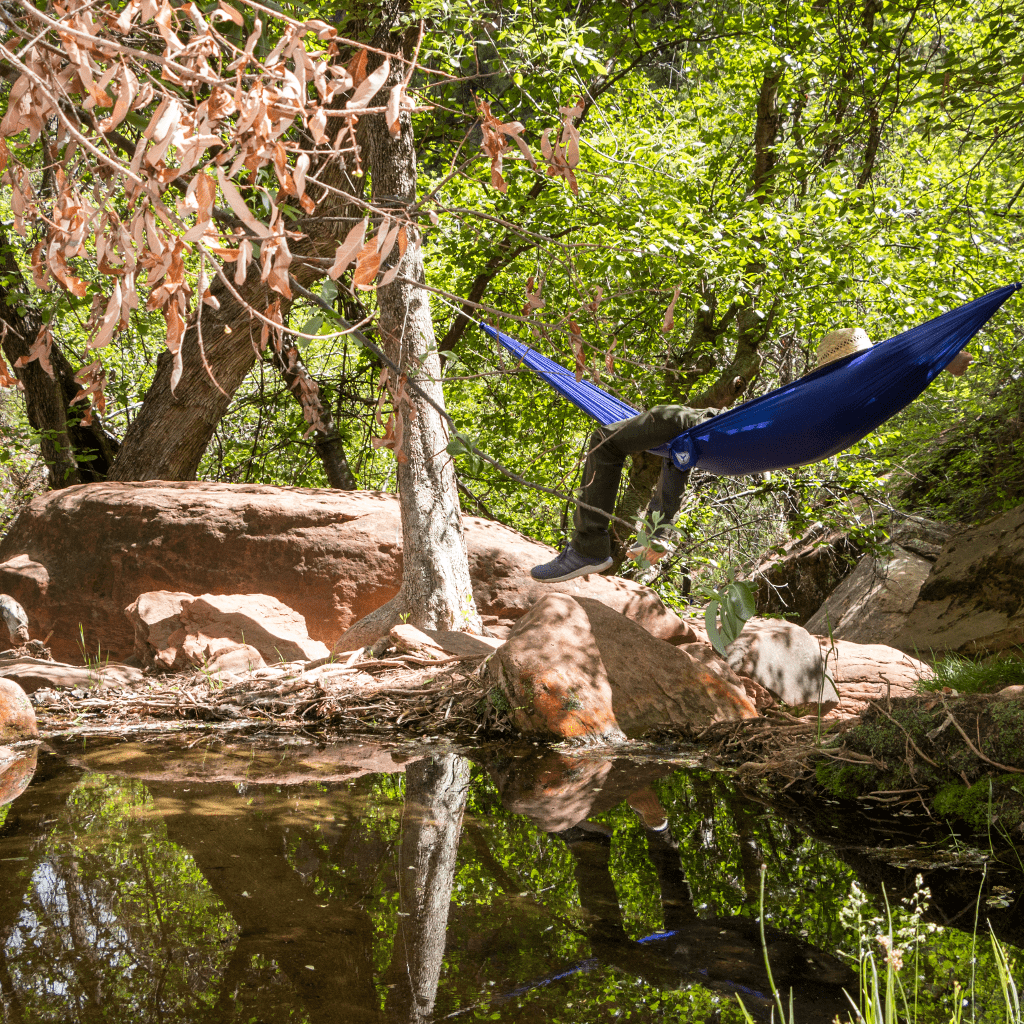 A blue Ultralight Hummingbird Hammocks Single Hammock, featuring parachute technology, suspended between trees over a rocky terrain with a small reflective water body beneath.