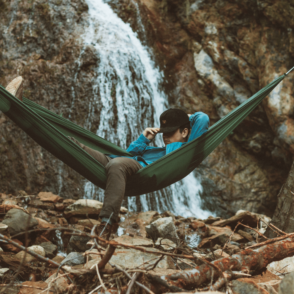 A person lounging in a Hummingbird Hammocks Ultralight Single Hammock by a waterfall, looking at their phone.