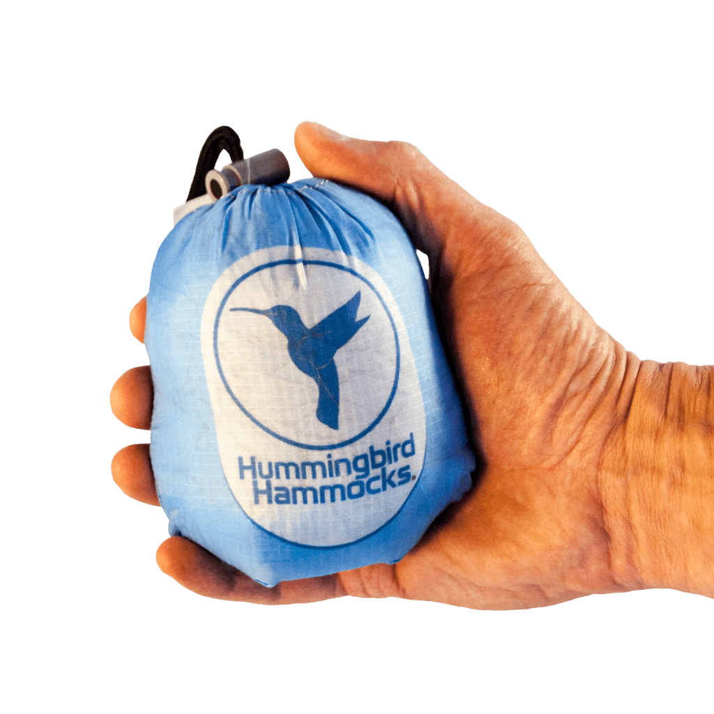 A hand holding a compact Hummingbird Hammocks Single Hammock bag featuring a Button Link attachment system in front of a green background.