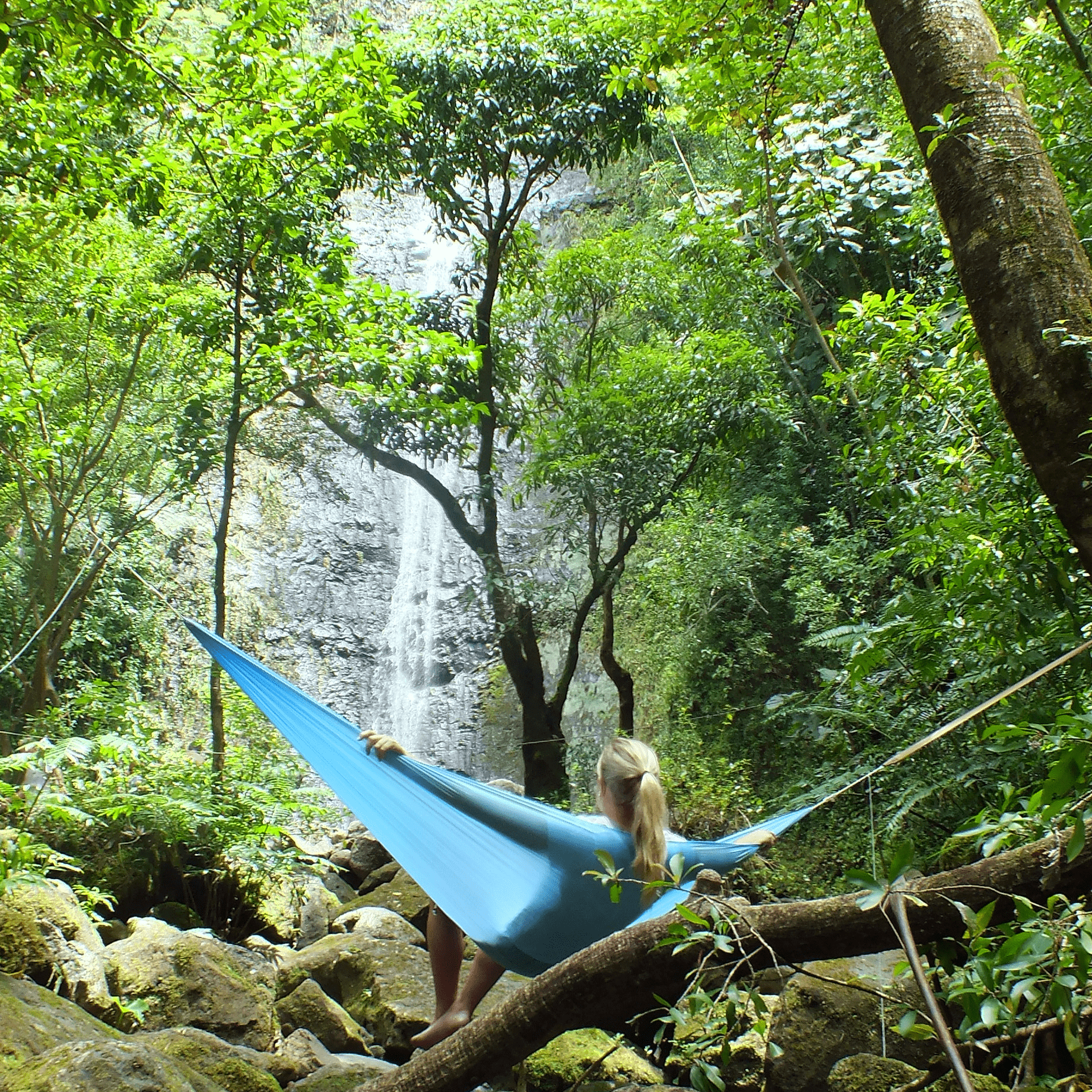 A woman relaxes in a Hummingbird Hammocks Ultralight Single Hammock in a lush forest, facing a large waterfall surrounded by rocks and greenery.
