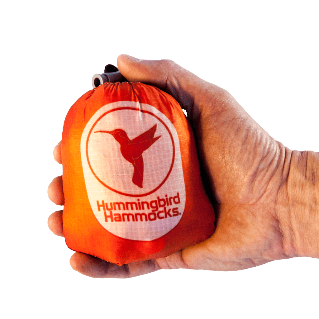 Hand holding a small, orange stuff sack labeled "Hummingbird Hammocks" with a white hummingbird logo, featuring an Ultralight Single Hammock from Hummingbird Hammocks, against a green background.