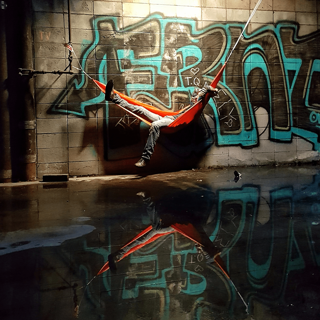 A person with jeans and sneakers swinging in a Hummingbird Hammocks Ultralight Single Hammock in a dimly lit room with graffiti on the walls and a reflective floor.