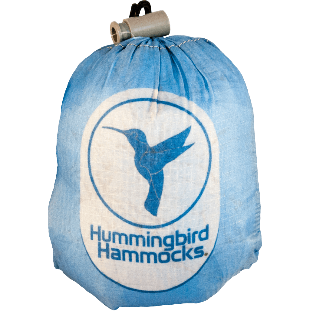 A blue portable bag labeled "Hummingbird Hammocks Single Hammock" using the Button Link attachment system, with a white logo featuring a hummingbird in flight.