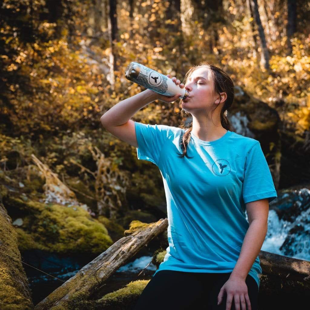 A woman in a blue shirt drinks from a Hummingbird Hammocks Topographic Stainless Steel Water Bottle by a forest stream.