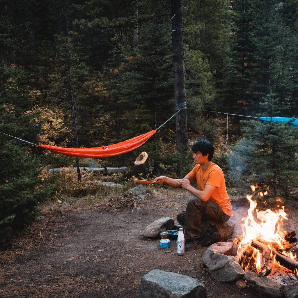 A person sitting by a campfire, tossing a pancake in a skillet, next to a hammock strung between trees in a forest, with a Topographic Stainless Steel Water Bottle by Hummingbird Hammocks beside them.