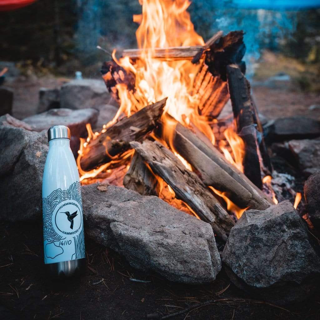 A reusable Topographic Stainless Steel Water Bottle by Hummingbird Hammocks with a bird design stands in front of a campfire surrounded by rocks.