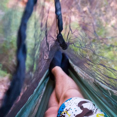 View from inside a hammock with Martin Bug Net - Bargain Bin, showing a person's legs and colorful patterned shorts, surrounded by a forest. (Hummingbird Hammocks)