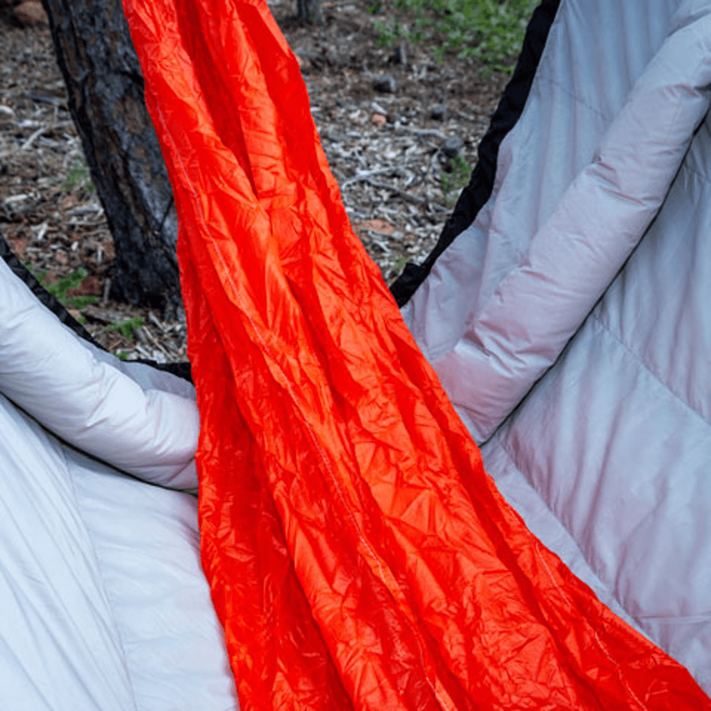 Close-up of a bright orange Puffin Underquilt by Hummingbird Hammocks on a gray sleeping pad, partially unzipped, located in a forest setting.