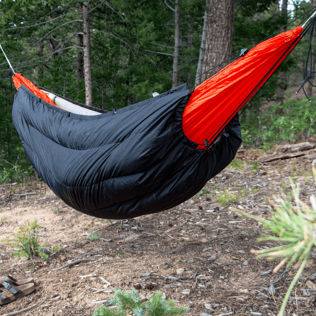 A red and black Hummingbird Hammocks Puffin Underquilt hangs between trees in a forested area.