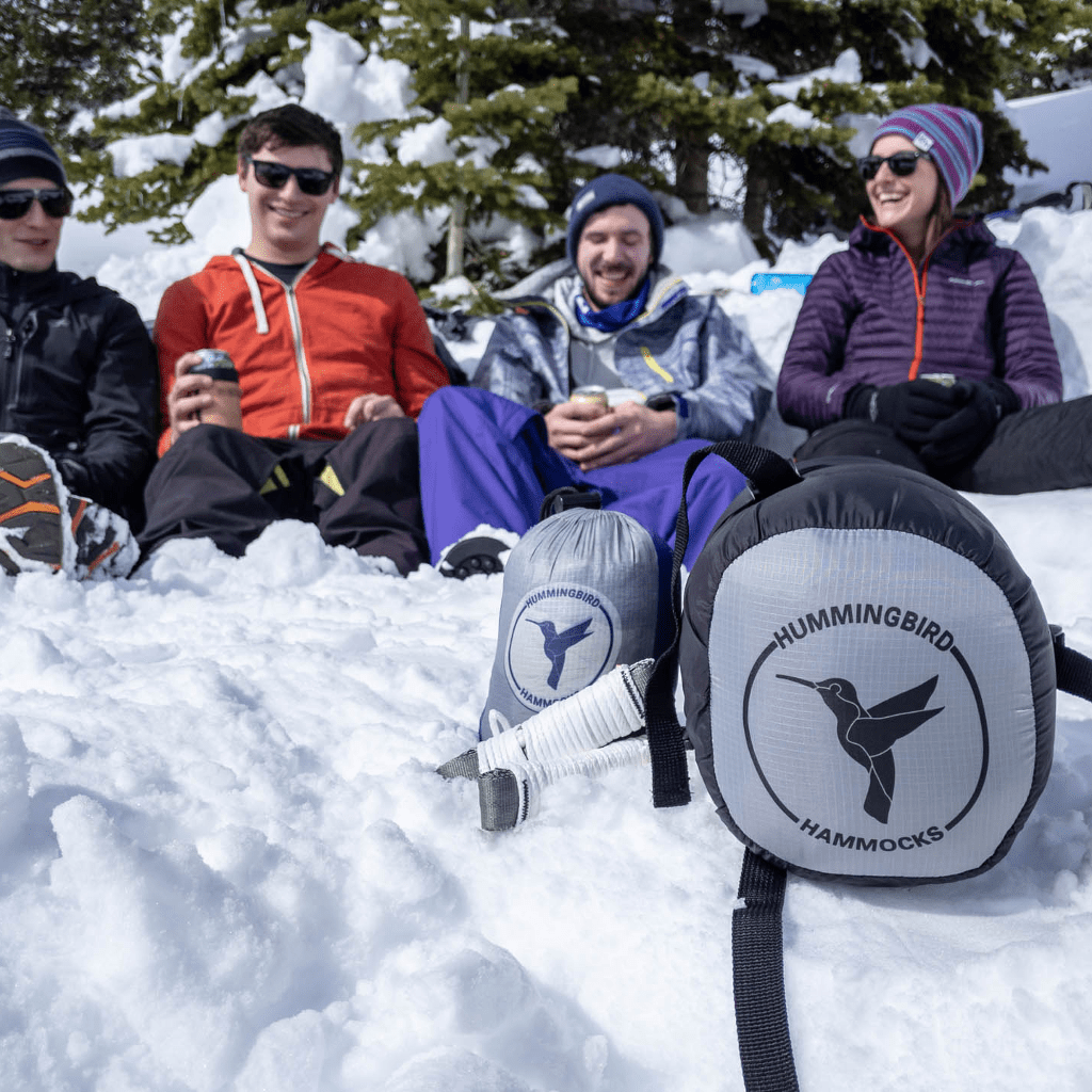 Four friends smiling and sitting in snow, with two Hummingbird Hammocks Puffin Underquilts featuring an advanced suspension system in the foreground, wearing winter gear.