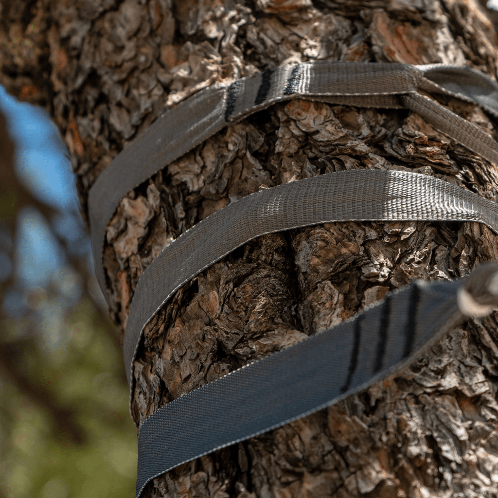 Ultralight gray Hummingbird Hammocks Tree Straps+ wrapped around a tree trunk, used for securing or supporting objects, against a blurred background of tree bark.