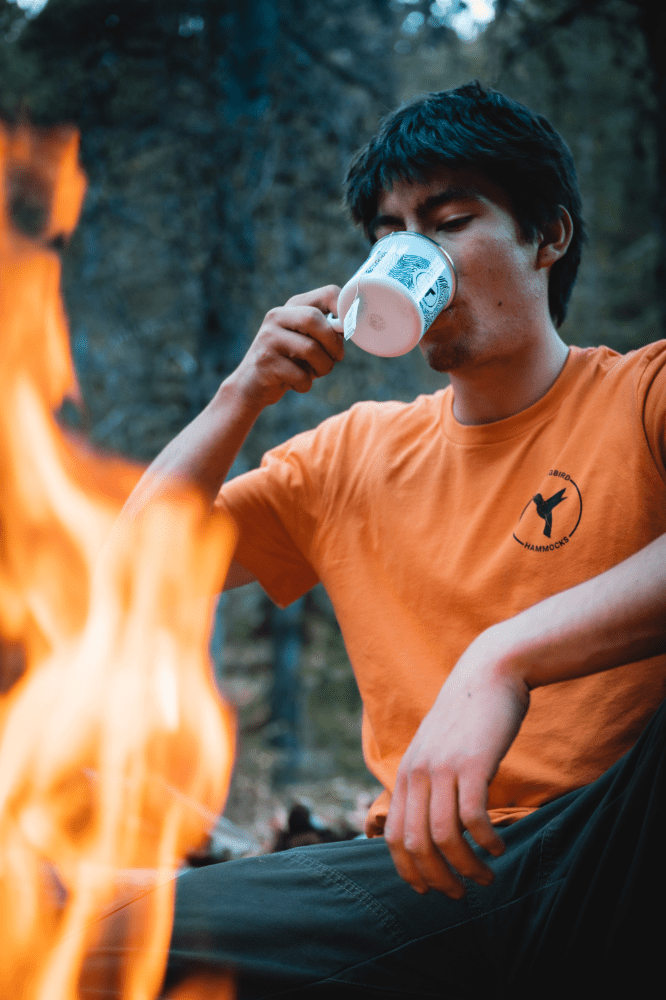A man in an orange T-shirt drinking from a Topo Enamel Mug by Hummingbird Hammocks beside a campfire.