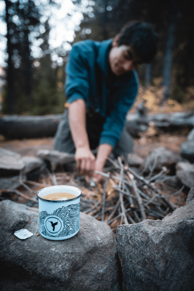A person preparing a campfire in a forest setting, with a focus on a Topo Enamel Mug by Hummingbird Hammocks placed on a rock in the foreground.